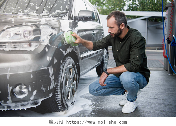 一个男人在用泡沫清洗车辆英俊的人蹲和清洁汽车清洗与抹布和泡沫